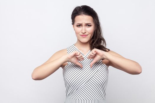 Portrait of sad young brunette woman with makeup and striped dress standing, holding thumbs down and looking at camera with sadness and dislike. indoor studio shot, isolated on grey background.
