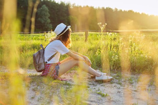 Happy teenager girl in hat with backpack sitting on country road, enjoying summer vacation, picturesque nature, copy space