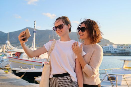 Happy family on vacation, mom and teenage daughter taking selfie on smartphone, sea bay with yachts summer sunset background