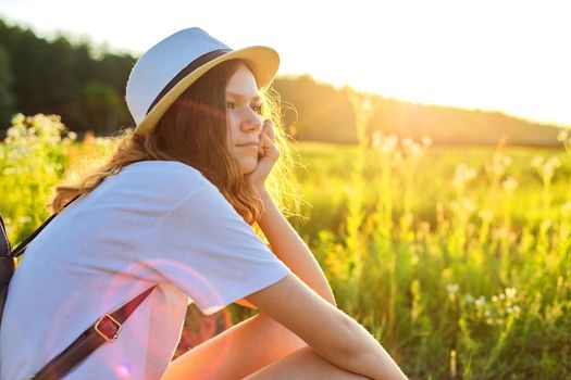 Happy teenager girl in hat with backpack sitting on country road, enjoying summer vacation, picturesque nature, copy space