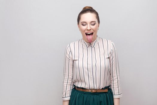 portrait of funny crazy beautiful young woman in striped shirt with makeup and collected ban hairstyle, standing with closed eyes and tongue out. indoor studio shot, isolated on grey background.