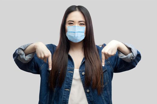 Here and right now. Portrait of serious bossy brunette asian young woman with surgical medical mask in blue denim jacket standing and pointing down. studio shot, isolated on grey background.