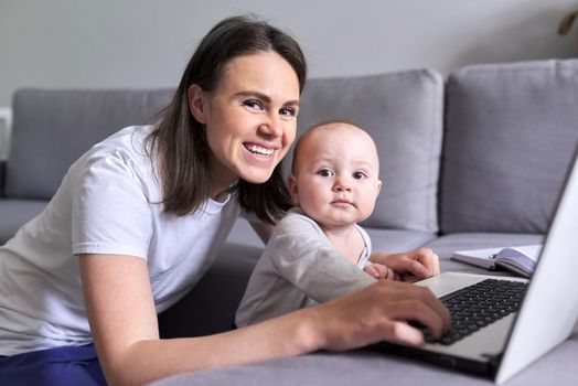 Young smiling mom with toddler baby look together at laptop monitor sitting at home on the couch