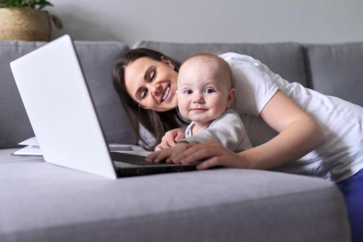 Young smiling mom with toddler baby look together at laptop monitor sitting at home on the couch