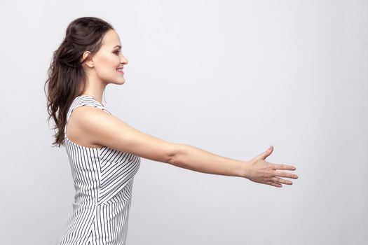 Profile side view portrait of happy beautiful young brunette woman with makeup and striped dress standing and giving hand to greeting and toothy smile. indoor studio shot, isolated on grey background.