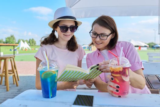 Happy mom and daughter teenager together in a summer outdoor cafe talking, drink soft drinks, look at a notebook