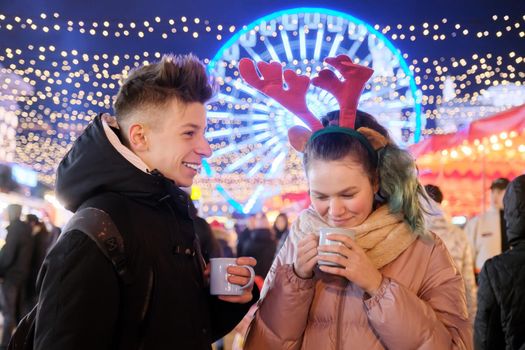 Christmas time, New Year holidays. Young people, couple of teenagers having fun at Christmas market, drinking hot tea from mugs, talking, laughing, background garlands of evening city, Ferris wheel