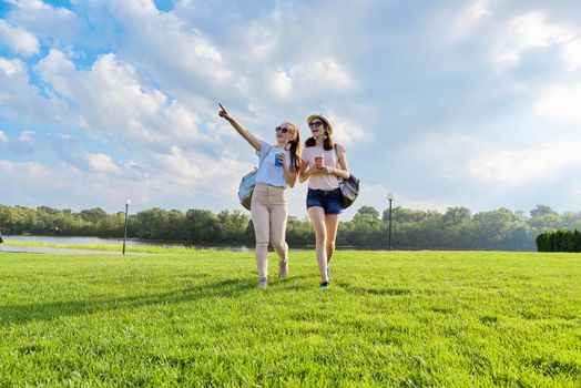 Two teenage girls walking together on the lawn with drinks on a sunny summer day, green grass and blue sky in the clouds background