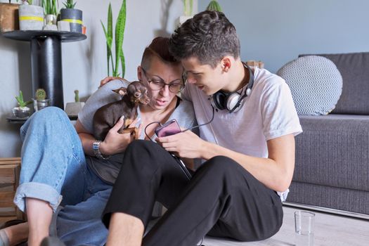 Happy talking mom and teenage son, parent and child sitting at home on the floor with pet little dog toy terrier, real friendly loving family