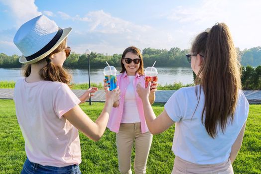 Portrait of happy mother and daughters teenagers 14 and 16 years old, girls with summer drinks. Background nature, park, river