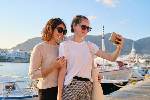 Happy family on vacation, mom and teenage daughter taking selfie on smartphone, sea bay with yachts summer sunset background