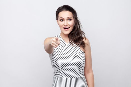 Portrait of amazed happy beautiful young brunette woman with makeup and striped dress standing, looking and pointing at camera with surprised face. indoor studio shot, isolated on grey background.
