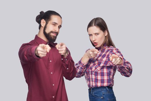 Hey you. Portrait of serious bearded man with black collected hair and woman in casual style standing and pointing at camera with serious face. indoor studio shot, isolated on gray background.