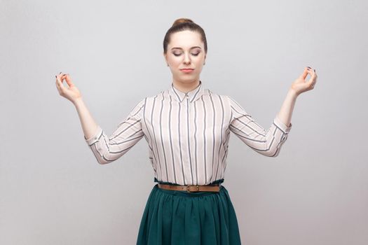 Portrait of meditating beautiful young woman in striped shirt and green skirt and collected ban hairstyle, standing in yoga pose with closed eyes. indoor studio shot, isolated on grey background.