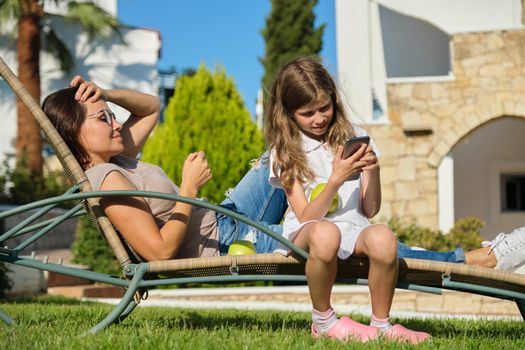 Mom and daughter child rest together sitting in an outdoor chair on the lawn, girl having fun with smartphone
