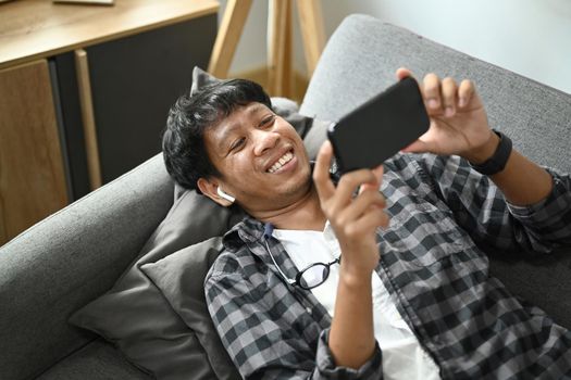 Happy young asian man lying on comfortable couch and using smart phone.