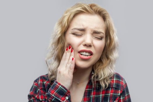 Tooth ache. Portrait of beautiful blonde young woman in casual red checkered shirt standing and touching her painful teeth with closed eyes. indoor studio shot, isolated on grey background.