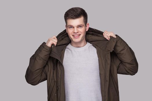 Portrait of young happy handsome man standing in light gray shirt and dark green parka looking at camera with toothy smile. indoor studio shot, isolated on gray background.