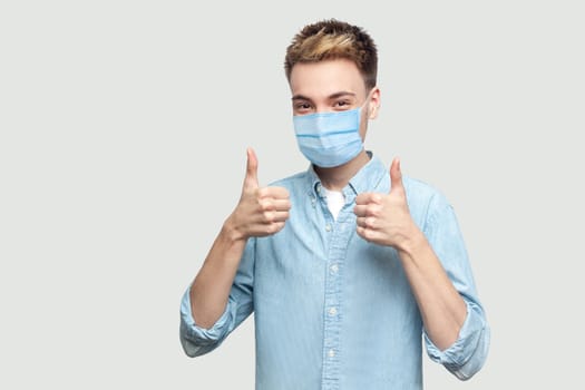 Portrait of happy satisfied successful handsome young man with surgical medical mask in light blue shirt standing, thumbs up and looking at camera. indoor studio shot on grey background copy space.