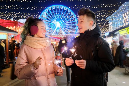 Christmas and New Year holidays, happy teenage couple with sparklers celebrating and having fun at Christmas market, sparkling lights of garlands of evening city ferris wheel background
