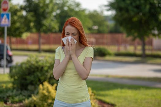 Young redhair woman sneezing in summer in front of blooming tree. Pollen Allergy symptoms