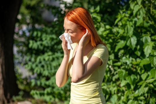 Young redhair woman sneezing in front of blooming tree. Pollen Allergy symptoms, green park in background