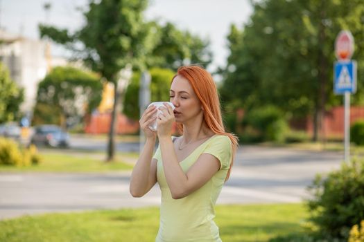 Young redhair woman sneezing in spring in front of blooming tree. Pollen Allergy symptoms