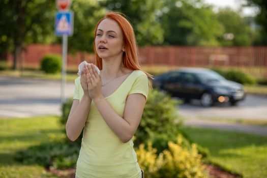 Young redhair woman sneezing in summer in front of blooming tree. Pollen Allergy symptoms