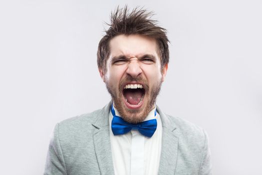 Closeup portrait of anger crazy handsome bearded man in casual grey suit and blue bow tie standing shouting or screaming with closed eyes. indoor studio shot, isolated on light grey background.