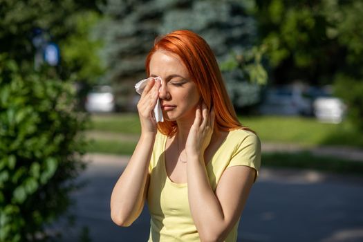 Young redhair woman sneezing in front of blooming tree. Pollen Allergy symptoms, green park in background
