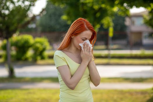 Young redhair woman sneezing in front of blooming tree. Pollen Allergy symptoms