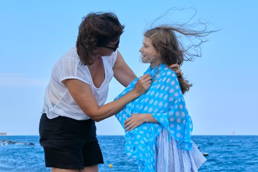 Happy mother and daughter walking together along sea beach, mom cares for her kid. Family weekend, travel, summer vacation concept