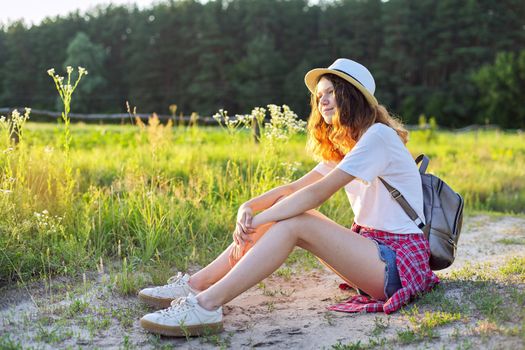 Happy teenager girl in hat with backpack sitting on country road, enjoying summer vacation, picturesque nature, copy space