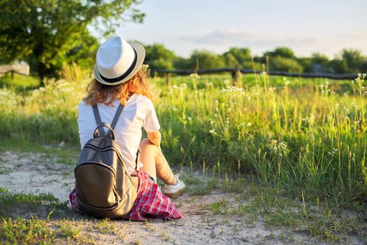 Happy teenager girl in hat with backpack sitting on country road, enjoying summer vacation, picturesque nature, copy space