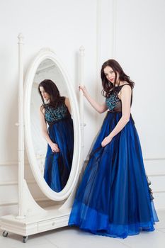 Gorgeous brunette woman in fashionable dress posing near big mirror at white room and looking at camera and smiling. indoor studio shot.