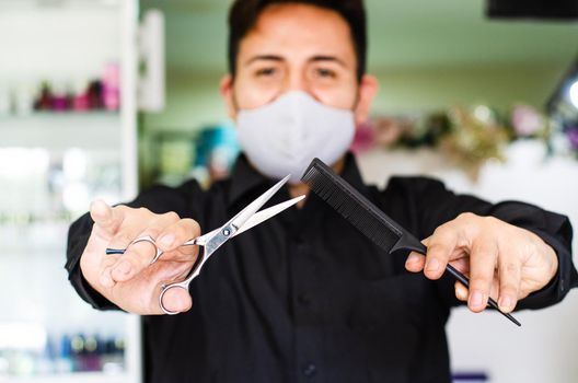 Portrait of an attractive barber, guy with safety mask, holding scissors and a comb in his hands, social distancing, preventive measures against the virus in pandemic