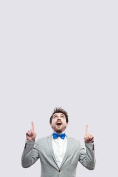 Portrait of amazed handsome bearded man in casual grey suit and blue bow tie standing looking and pointing at up copyspace. studio shot, isolated on light grey background.