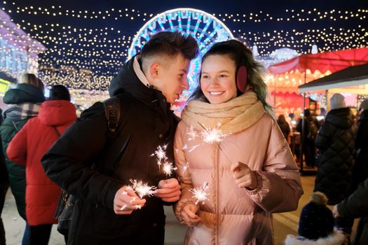 Christmas and New Year holidays, happy teenage couple with sparklers celebrating and having fun at Christmas market, sparkling lights of garlands of evening city ferris wheel background