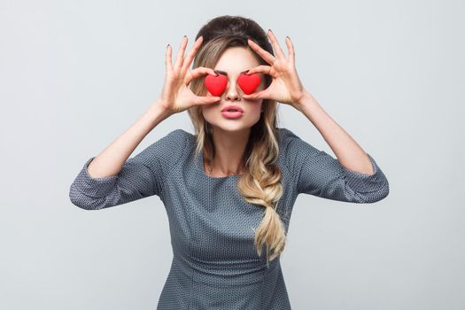 Loving eyes. Beautiful romantic caucasian young woman holding two valentine hearts in front of her eyes like glasses while standing against grey background. Copy space, indoor, studio shot, isolated
