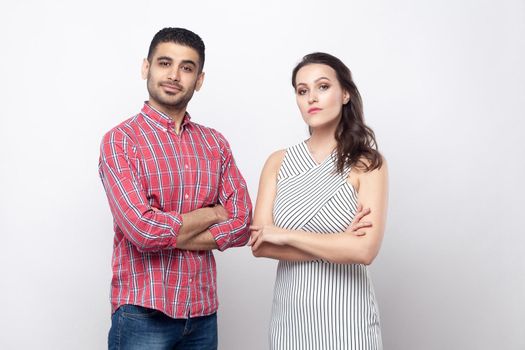 Portrait of serious proud handsome man in red checkered shirt and beautiful woman in white striped dress standing and looking at camera with crossed arms. indoor studio shot on grey background.