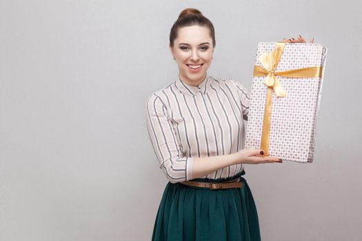 Kindness attractive young adult woman in white with green lines skirt standing and showing you present with yellow bow and toothy smile, looking at camera. Indoor,isolated,studio shot, gray background