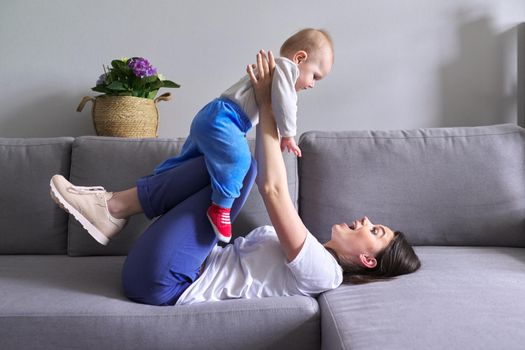 Happy mother with toddler son at home. Laughing woman playing with baby lying on sofa. Family, childhood, love, motherhood happiness concept