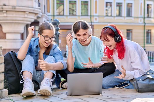 Teenage friends in headphones looking at laptop. Group of young people having fun laughing looking at laptop screen, sitting on sidewalk, urban style background. Youth, fun, lifestyle, leisure concept