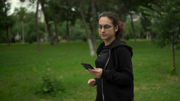 A young woman is jogging with her phone in the park. Asian girl with glasses running with app in phone move to earn. 4k