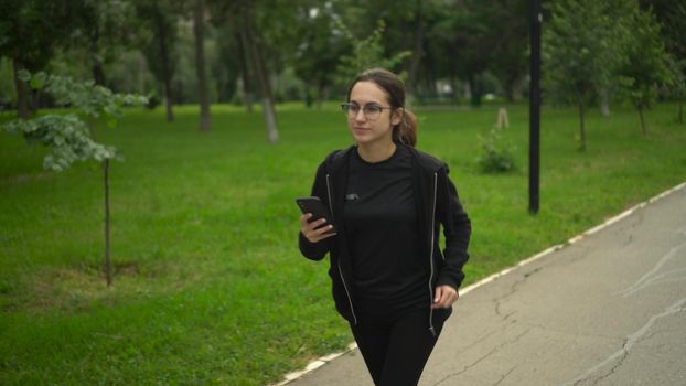 A young woman is jogging with a phone in her hand and rejoices. Asian girl with glasses is running with the application in the phone move to earn. 4k