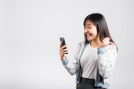 Woman excited holding mobile phone funky successful raise fists say yes! studio shot isolated white background, happy young female smiling surprised make winner gesture on smartphone