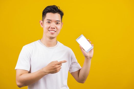 Happy Asian handsome young man smiling positive showing smartphone blank screen and pointing finger to screen, studio shot isolated on yellow background, making successful expression gesture concept