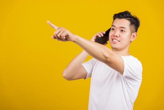 Asian handsome young man smiling positive showing smartphone blank screen and pointing finger to side away, studio shot isolated on yellow background, making successful expression gesture concept