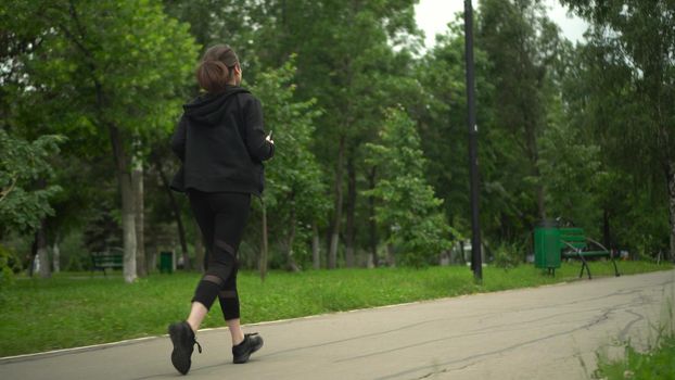 A young woman is jogging with her phone in the park. Asian girl with glasses running with app in phone move to earn. 4k