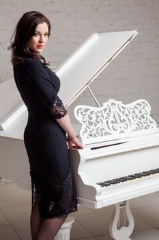 Side view profile of sensual brunette woman in lace black classic dress standing near white piano and looking at camera. indoor studio shot on white brick wall.